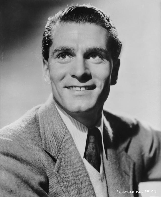 laurence olivier smiles and look upward past the camera, he wears a suit jacket, collared shirt and tie