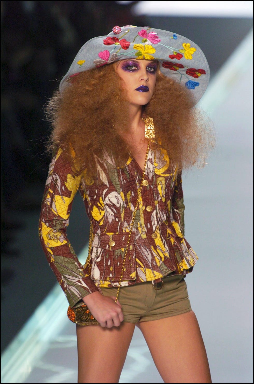 riley keough poses with her hand on a small purse while standing on a runway, she wears a hat with embroidered flowers, a floral patterned jacket, and khaki shorts