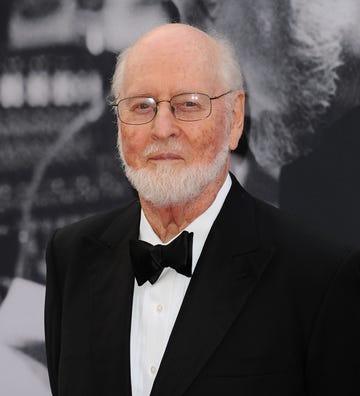 john williams smiles at the camera, he wears a black tuxedo with a white collared shirt and metal rimmed glasses