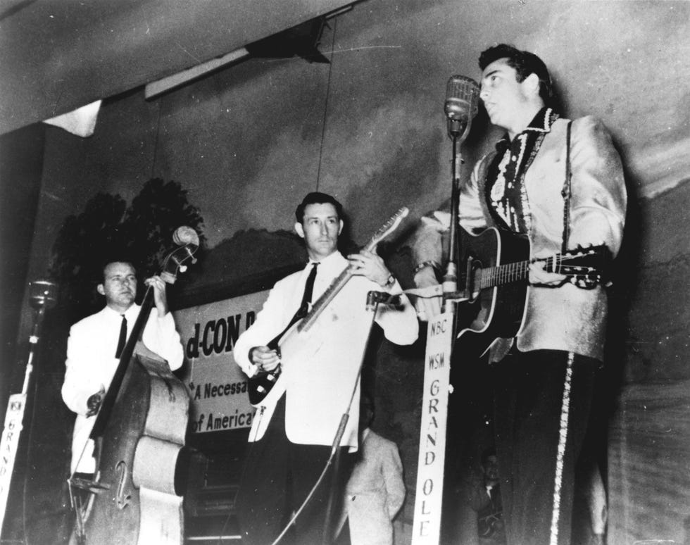 marshall grant, luther perkins, and johnny cash perform on a stage, each wears a suit jacket, tie and dark pants, cash sings into a microphone on a stand