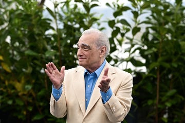 martin scorsese claps his hands while standing in front of greenery, he looks to the left and wears a tan suit jacket over a blue collared shirt