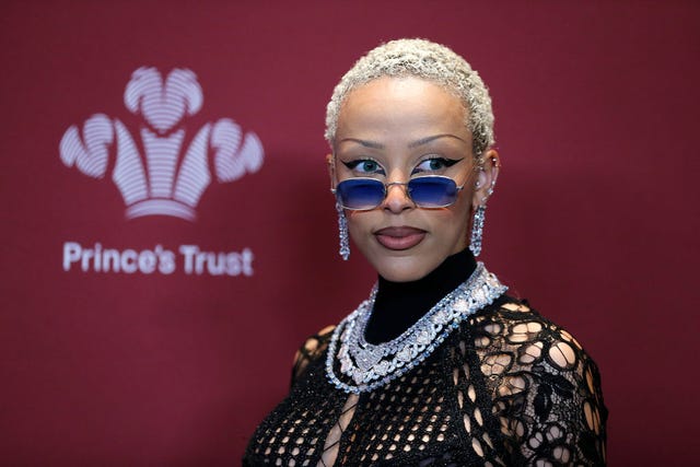 doja cat looks to the right, she wears a black lace top with diamond and silver jewelry and blue tinted glasses