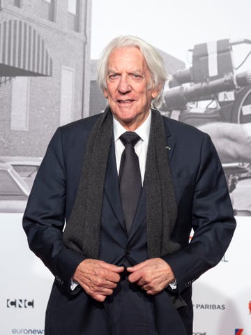 donald sutherland smiles at the camera, he wears a dark suit jacket and tie with a white dress shirt and a gray scarf