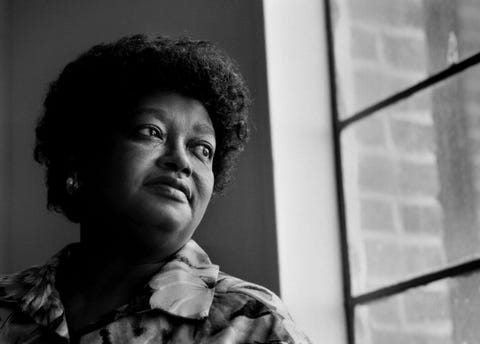 claudette colvin looks out a window, she wears a floral patterned collared shirt and an earring