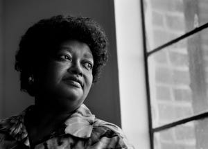 claudette colvin looks out a window, she wears a floral patterned collared shirt and an earring