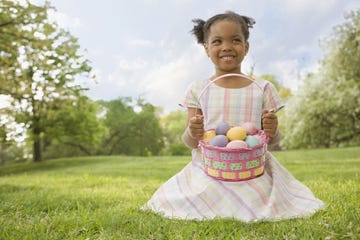 little girl smiling in green field wearing pastel plaid dress with basket full of colorful eggs found during easter egg hunt