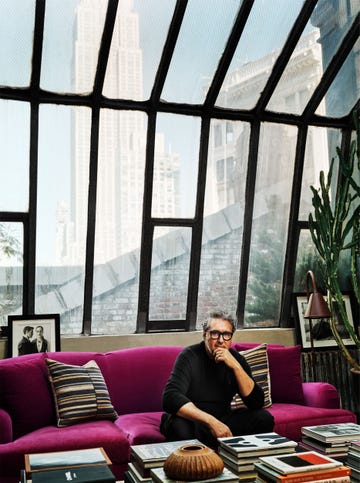 a person sitting on a bright purple couch in a modern interior with large studio windows showing the empire state building and a table stacked with books in front of him