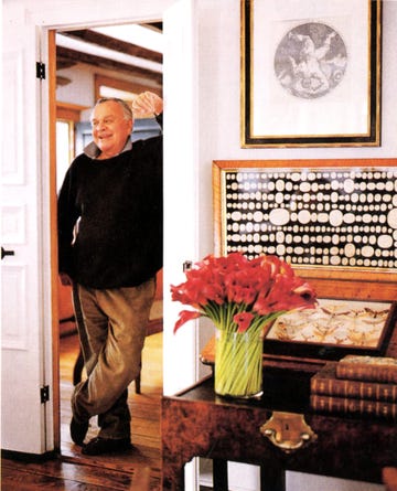 older man in a black sweater and baggy corduroys leans against a doorway with a small table in the foreground with a vase of red flowers and some old books on it