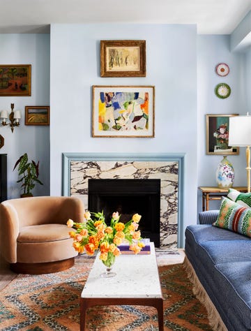 living room with light blue walls, square marble fireplace, light rust swivel chair, white marble topped cocktail table, blue sofa with accent pillows and bottom fringe, moroccan rug, colorful vase, multiple framed artworks