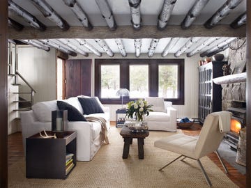 cozy living room with rustic white pine ceiling beams and modern furniture and a stone fireplace and a three panel window at the back