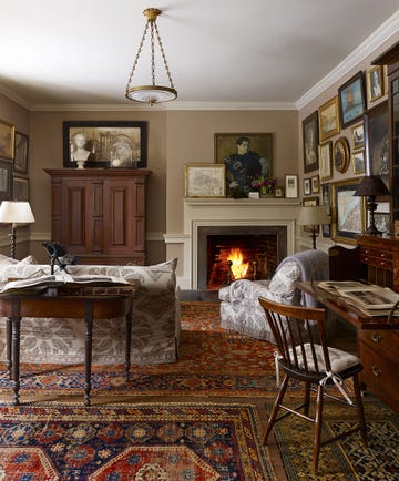 cozy room with several colorful rugs and a lit fireplace at the far end with paintings and framed prints around the wall and a windsor chair at a desk and an armoire and a sofa with a demilune table behind it