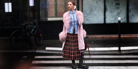 individual wearing a pink fur coat over a striped shirt and plaid skirt standing on a crosswalk