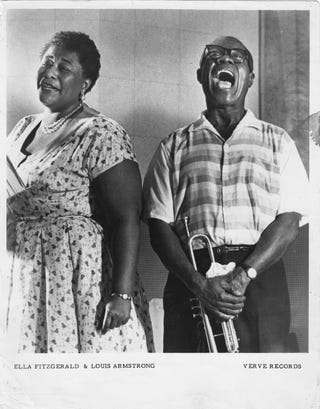 ella fitzgerald smiles and looks at the camera while wearing a short sleeve patterned dress, louis armstrong stands to the right with a wide mouth smile as he holds his trumpet, he wears a short sleeve button up shirt with pants and thick framed glasses