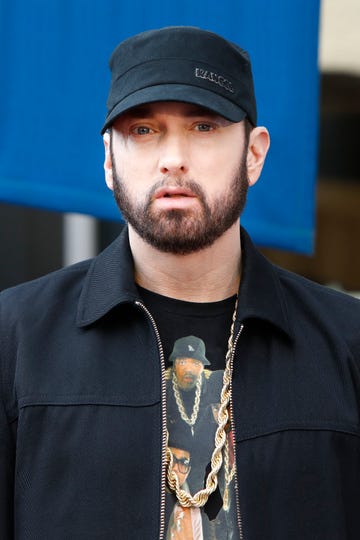 eminem looks at the camera with a straight face, he wears a black cap and jacket with a black graphic t shirt and golden chain necklace