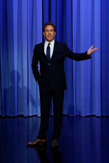 jerry seinfeld stands in front of a blue curtain and extends one arm out, he wears a dark suit with a white collared shirt and tie