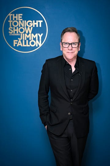 kiefer sutherland smiles at the camera, he wears black glasses, a black suit jacket and a black collared button up shirt