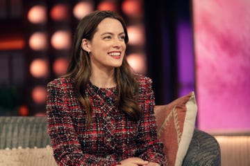 riley keough smiles and looks to the right as she sits on a couch, she wears a red, black and white tweed jacket