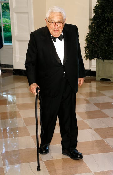 henry kissinger smiles at the camera, he wears a black suit with a black bowtie and a white collared shirt, he holds onto a cane while standing in a room