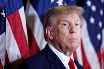 donald trump stands in front of several american flags that are out of focus in the background, he looks off camera to the right with a neutral expression on his face, he is wearing a navy blue suit jacket, white collared shirt and red tie