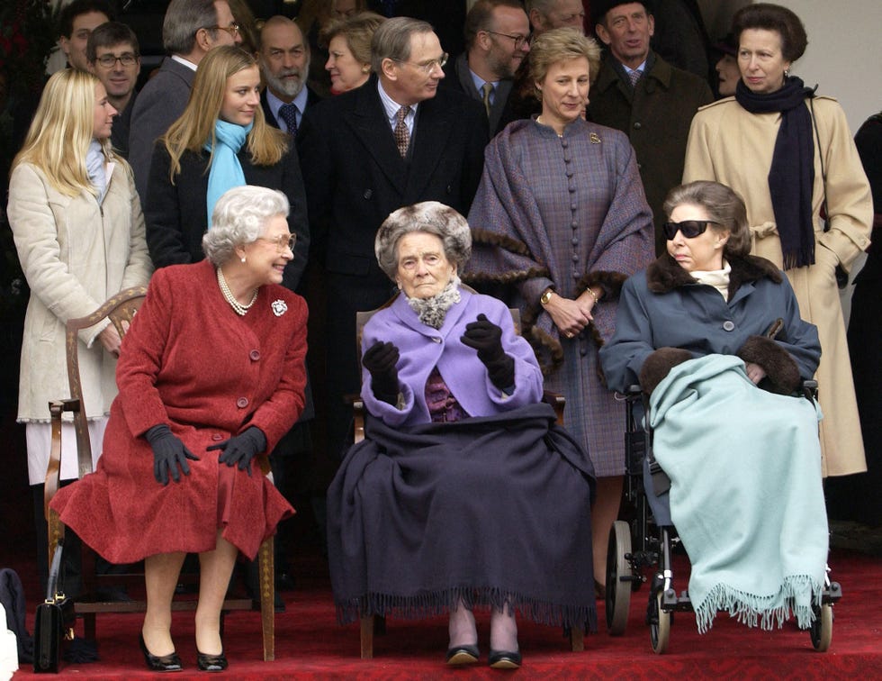 queen elizabeth, princess alice, and princess margaret sit on a red carpet as several people stand behind them, elizabeth wears a long red coat and black gloves, alice wears a purple jacket, black gloves, and fur hat with a dark blanket on her lap, margaret wears a blue fur trimmed coat with sunglasses and an ice blue blanket on her lap