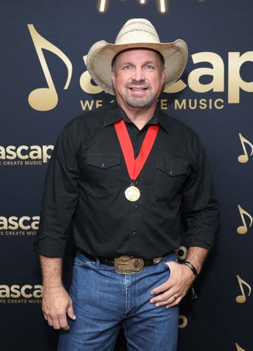 garth brooks smiles at the camera, he wears a straw cowboy hat, black collared shirt and a gold medal with a ribbon lanyard