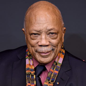 quincy jones smiles at the camera, he wears a dark suit jacket and matching tie with a pink collared shirt and multicolored band around his neck