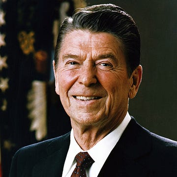 ronald reagan smiles at the camera, he wears a dark suit jacket, white collared shirt, and red patterned tie