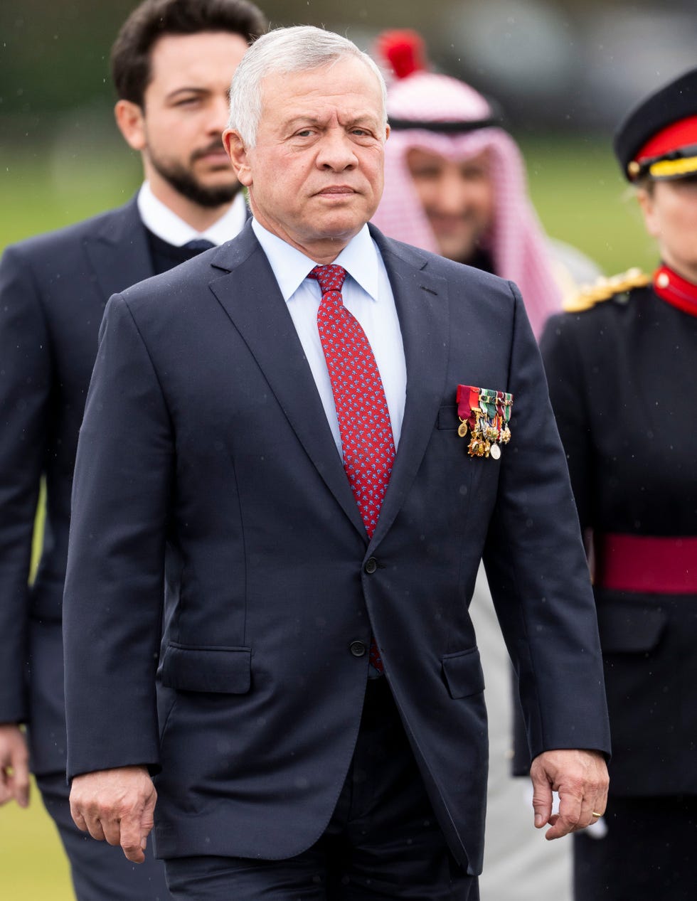 king abdullah ii of jordan stands and looks at the camera with a neutral expression, he wears a dark suit, white collared shirt and red tie with a blue pattern, his breast pocket is decorated with several medals