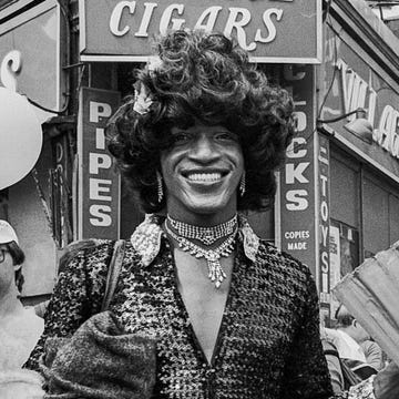 marsha p johnson in dark outfit and black hair smiles as she stands on the corner of christopher street and 7th avenue during the pride march