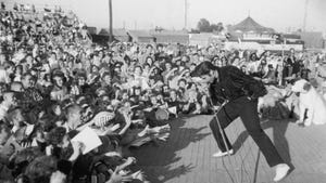 Elvis Presley performs outdoors on a small stage to the adulation of a young crowd.