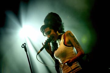 amy winehouse sings into a microphone she holds in one hand, she smiles and stands next to a microphone stand, she wears a white tank top