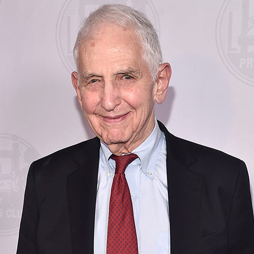 daniel ellsberg smiles at the camera, he is wearing a black suit jacket with a light blue collared shirt and red tie