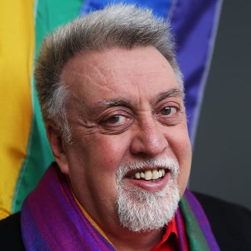 gilbert baker smiles at the camera, behind him is a rainbow flag and he wears a rainbow scarf with his black top