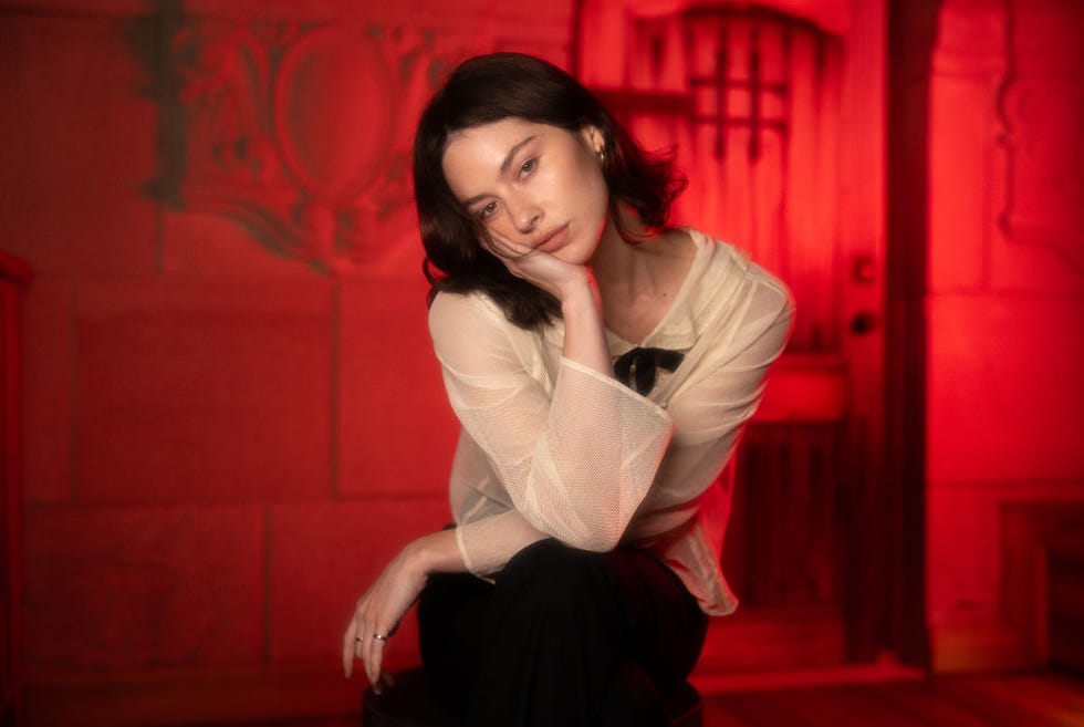 gracie abrams looks at the camera as she rests her face in one palm, she sits crosslegged and wears a white blouse with black pants