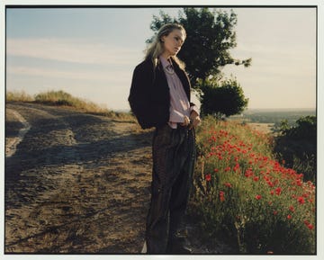 model stands on a dirt path lined by red flowers and overlooking a view wearing a lavender button up, a sweater, and trousers