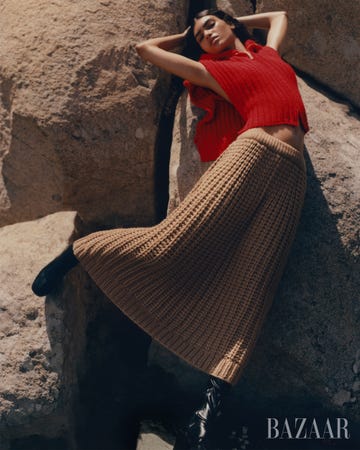 model leans against boulders in a red knit top and a camel knit skirt