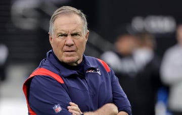 bill belichick looks angrily toward the camera with his arms crossed, he wears a blue new england patriots vest and matching shirt with an nfl logo on the right sleeve