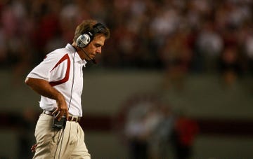 nick saban looks down and walks to the right, he wears a white polo shirt and khakis with a headset, he adjusts the headset receiver on his belt with on hand