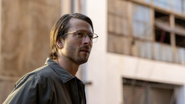 glen powell with long hair and glasses standing in front of an old warehouse in a scene from hit man