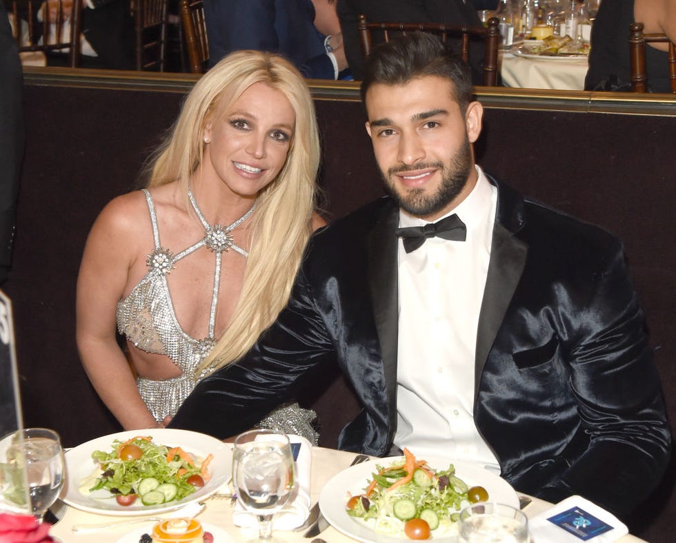 britney spears and sam asghari sit at a dinner table and smile at the camera, she wears a glittering silver dress with cutouts, he wears a black tuxedo and rests one arm across her lap under the table