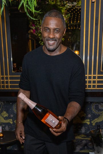 idris elba smiles at the camera, he wears a black shirt and flowers and lights are hanging from the ceiling behind him