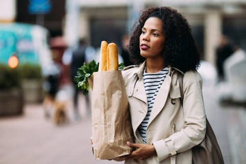 a stylish woman in a trench coat carrying a paper bag of groceries with baguettes and greenery in an urban area