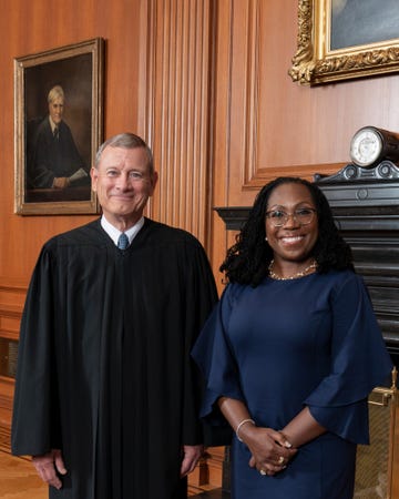 supreme court chief justice john roberts smiles at the camera, he is wearing his black justice robes with a blue tie, behind him is a wood paneled wall with a partial portrait in view