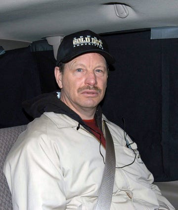 gary ridgeway looks at the camera while buckled into a car seat, he wears a dark ball cap, tan jacket and red shirt