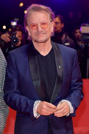 bono smiles and looks past the camera, he stands on a red carpet with photographers behind him, he wears a blue and black suit jacket with a black shirt, orange circular glasses, and earrings