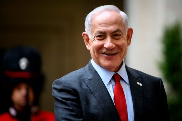 benjamin netanyahu smiles and looks to the left, he wears a gray suit jacket, light blue dress shirt, red tie and israel flag pin