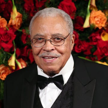 james earl jones smiles at the camera, he wears wire rimmed glasses and a black tuxedo with a vest and bowtie, behind him is a wall of flowers