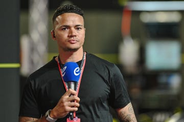 jermaine jenas, a man stands looking at the camera with a serious facial expression, he has short curly black hair and wears a black tshirt, he is standing in a sports arena holding a microphone