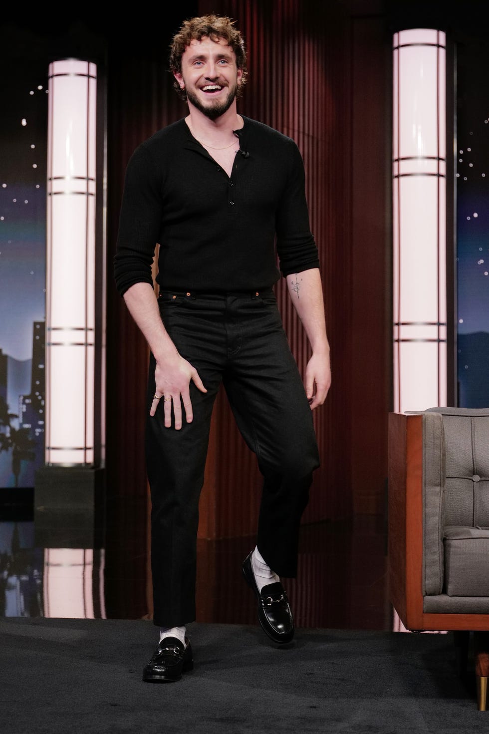 paul mescal smiles while walking in a tv studio, he wears a black v neck shirt and thin necklace