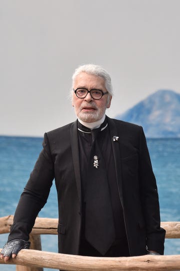 karl lagerfeld looks at the camera with a straight face, he wears a black suit with decorative pins on his black tie and jacket lapel, he also wears black framed aviator glasses